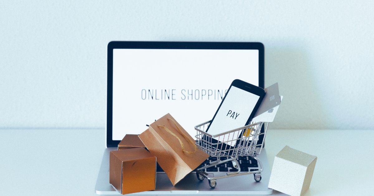 Um notebook aberto, escrito shopping online na tela com um carrinho de compras e pacotes espalhados ao redor e acima do teclado.