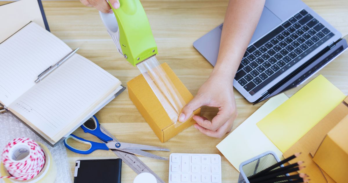 Pessoa embalando uma caixa de produto com notebook e materiais de e-commerce ao lado, representando um negócio de dropshipping