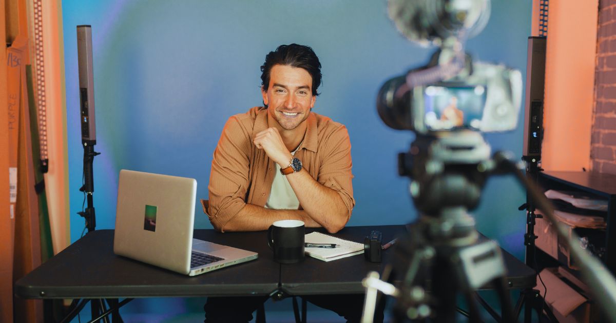 Rapaz sentado em uma mesa com estúdio atrás, sendo filmado por uma câmera enquanto produz conteúdo para marketing digital.
